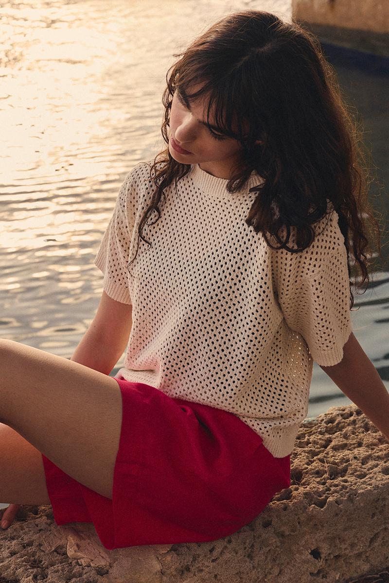 A woman wearing a cream crochet top and pink shorts from BITTE KAI RAND, sitting on a rocky ledge by the water during the golden hour.