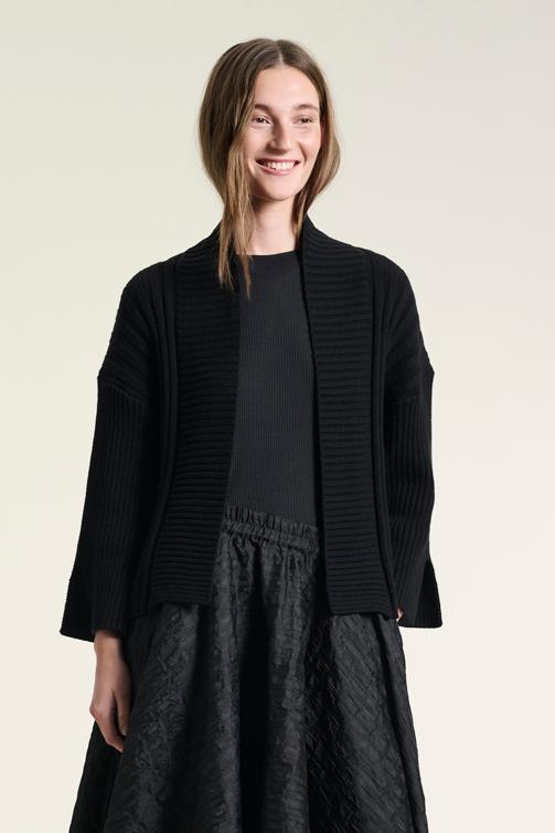 A smiling woman wearing a black BITTE KAI RAND cardigan with a ribbed texture, paired with a dark top and a textured skirt.